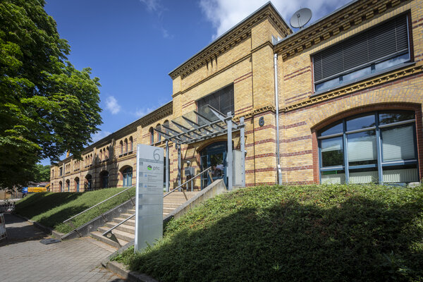 Gelbes Backsteingebäude mit zwei Etagen. Eine Treppe führt zum Haupteingang, links und rechts davon liegen Grünflächen.